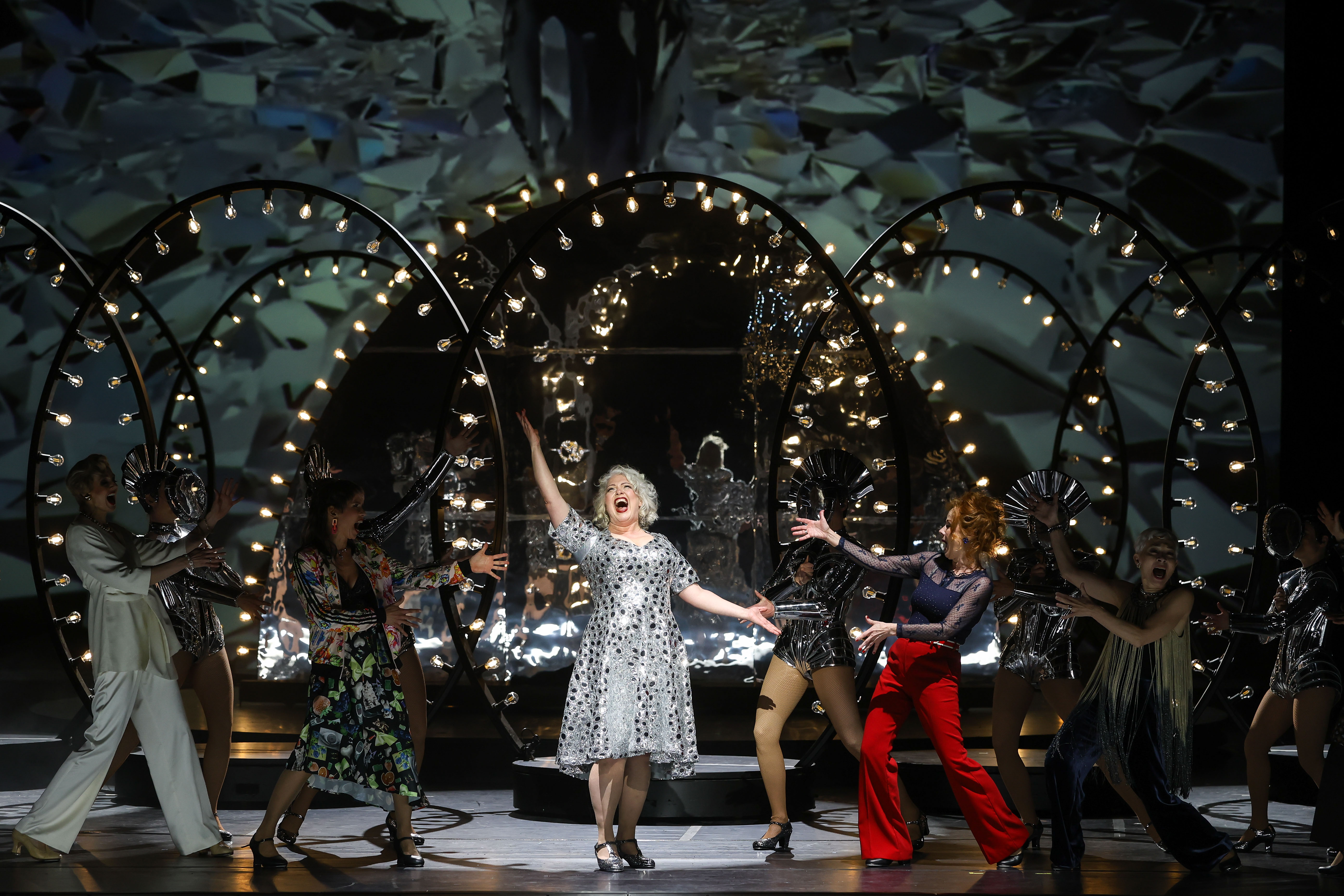 Szenefoto des Musicals "Follies" an der Volksoper wien, im Zentrum eine der Hauptdarstellerinnen in einem silbenen Kleid mit schwarzen Tupfen, umgeben von Ensemblemitgliedern