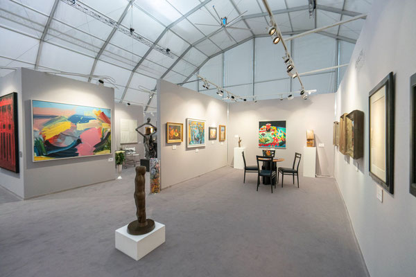 Exhibition view: Different paintings on light gray walls, in the foreground a small brown statue. The whole exhibition is located in a large tent