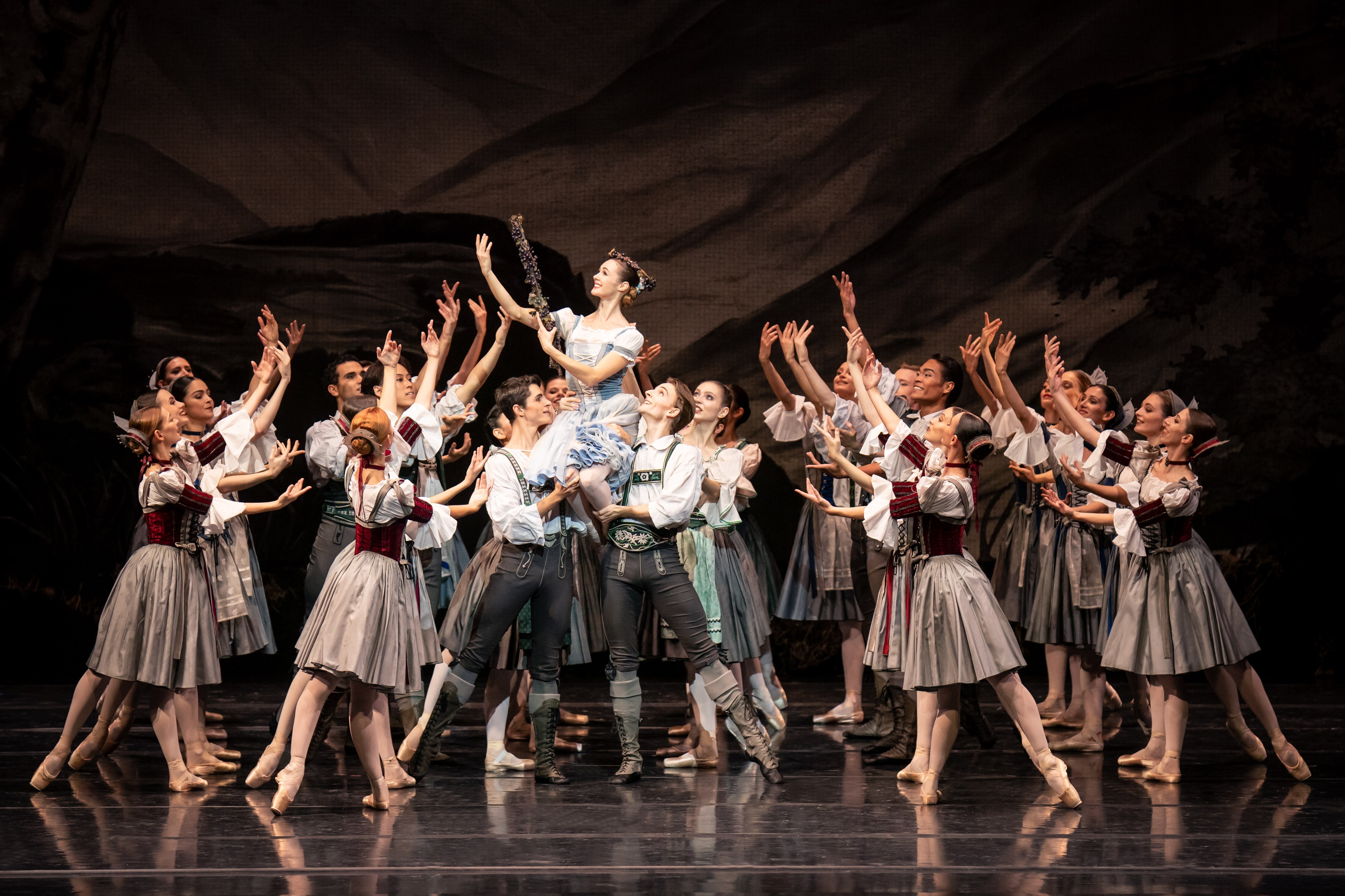 Gruppe von Tänzerinnen, die Frau tragen Drindl-Kostüme, die Männer Lederhosen mit weißem Hemd, die Ballerina in einem hellblauem Dirndl wird auf den Schultern von zwei Männern getragen