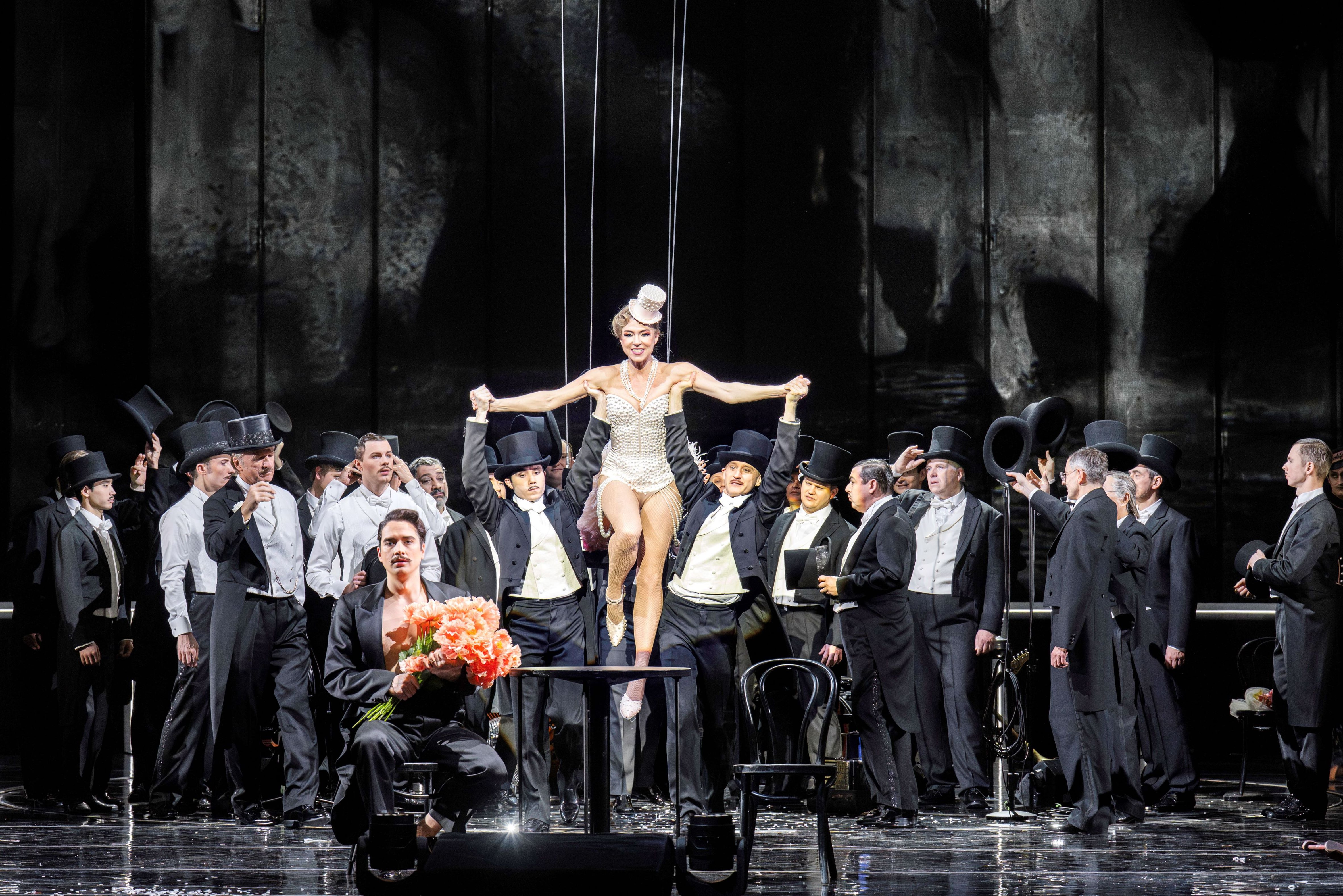 Das Foto zeigt die Hauptdarstellerin in weißem Perlentrikot mit weißem Hüttchen umgeben von einer Gruppe Männer in Frack und Zylinder, davor ein Mann mit einem rotorangen Blumenstrauß