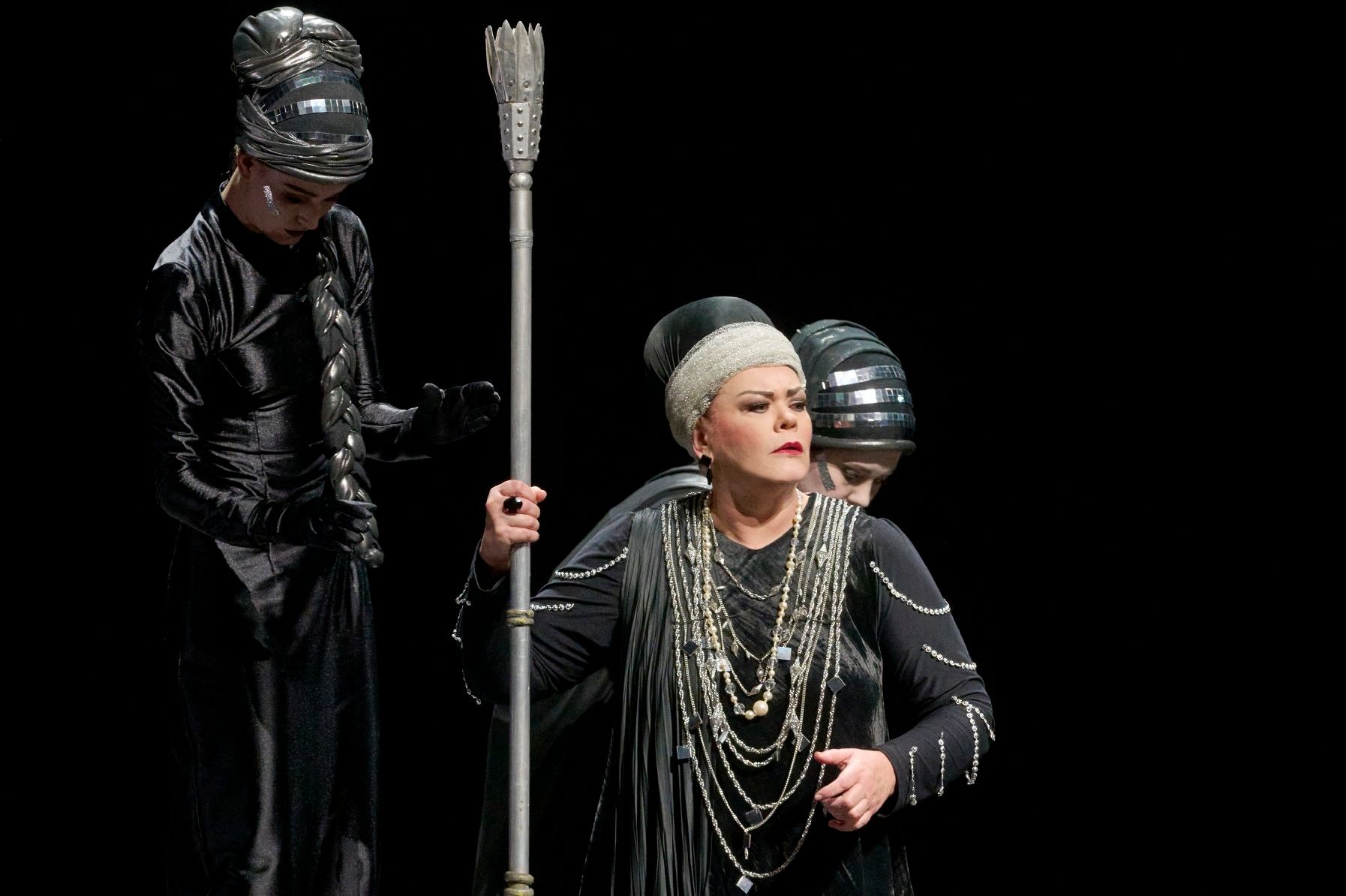 Das Szenefoto aus der Oper "Elektra" zeigt eine dominante Frau mit einer Lanze in der rechten Hand, sie trägt ein schwarzsilbernes Kleid mit vielen langen Halsketten und einer Art Turban auf dem Kopf. Hinter ihr zwei Personen in schwarzer Kleidung
