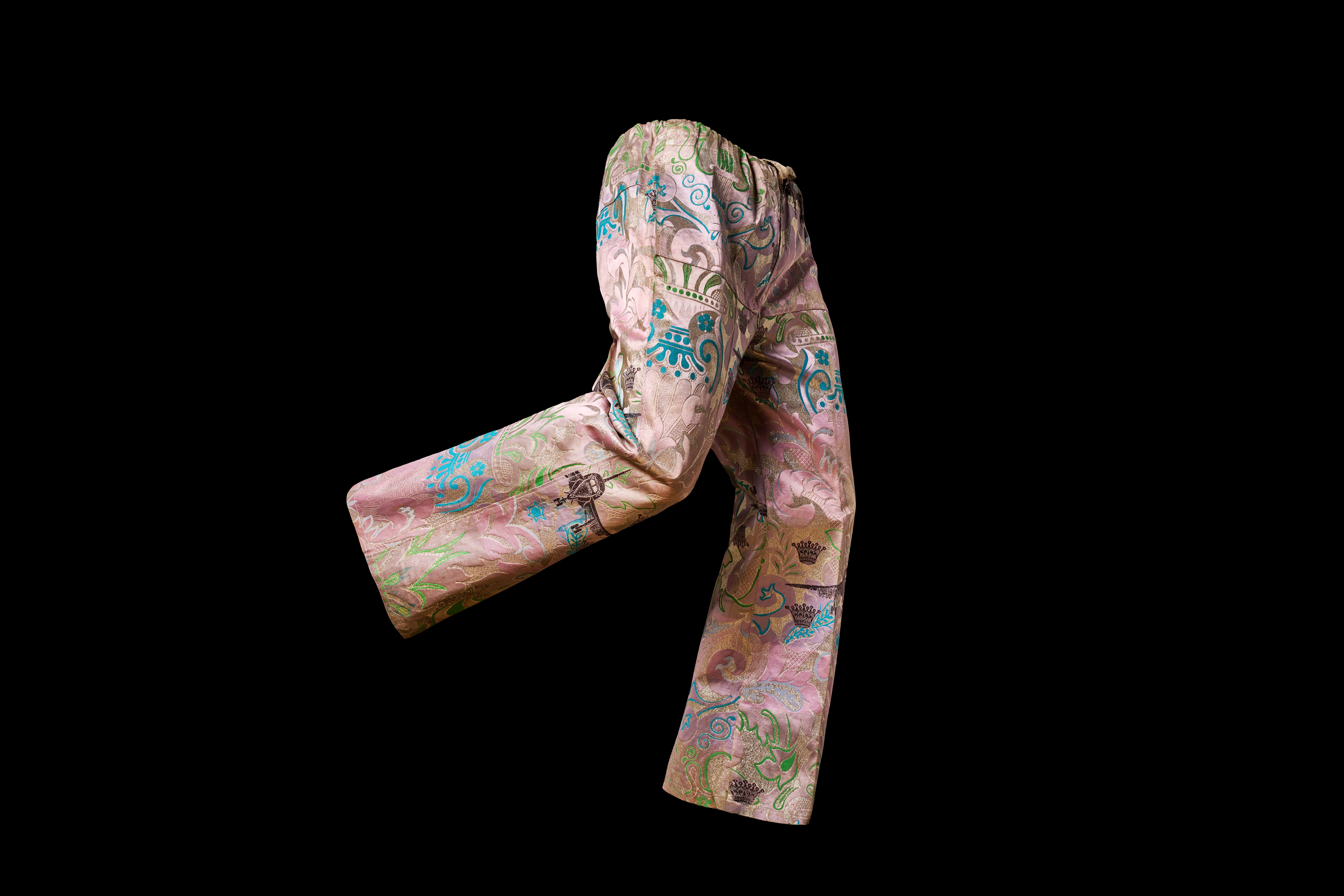 The photo shows a pair of trousers from Nigeria from around 1980, made of pale pink cotton fabric with colorful sewn-on ornaments in turquoise, brown, green and gold