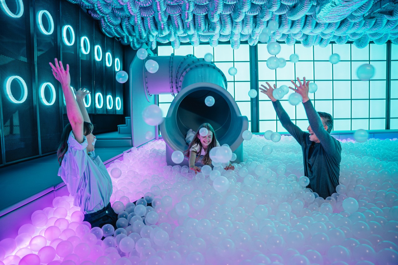 The photo shows a woman and a man having fun in a ball pool, a girl slips out of a tube into this ball pool, the scene is bathed in blue and magenta light at the edges