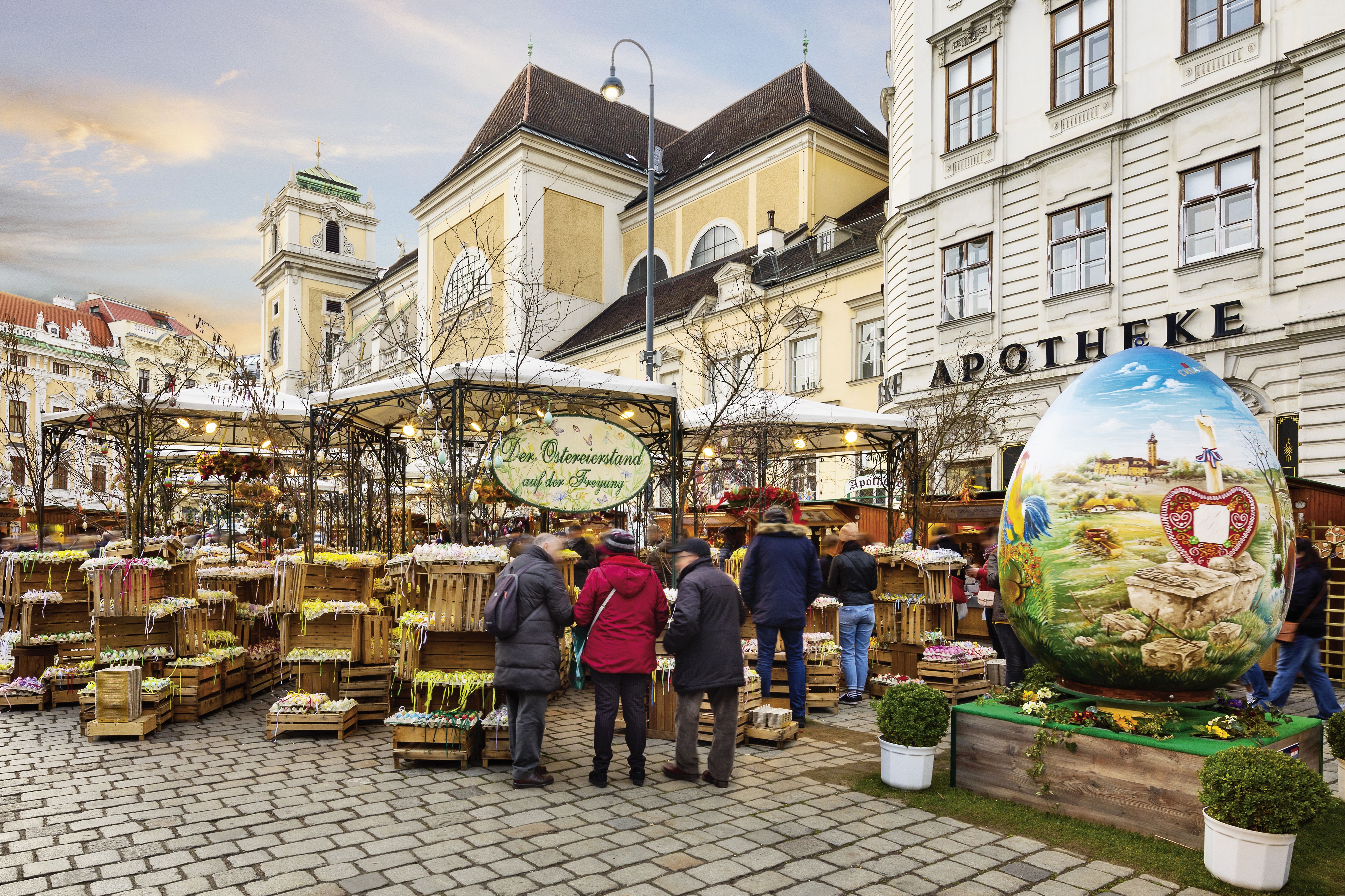 Szene auf dem Altwiener Ostermarkt mit Ständen, Kisten mit bemalten Ostereiern, im Vordergrund ein übergroßes bemaltes Ei, im Hintergrund eine Kirche