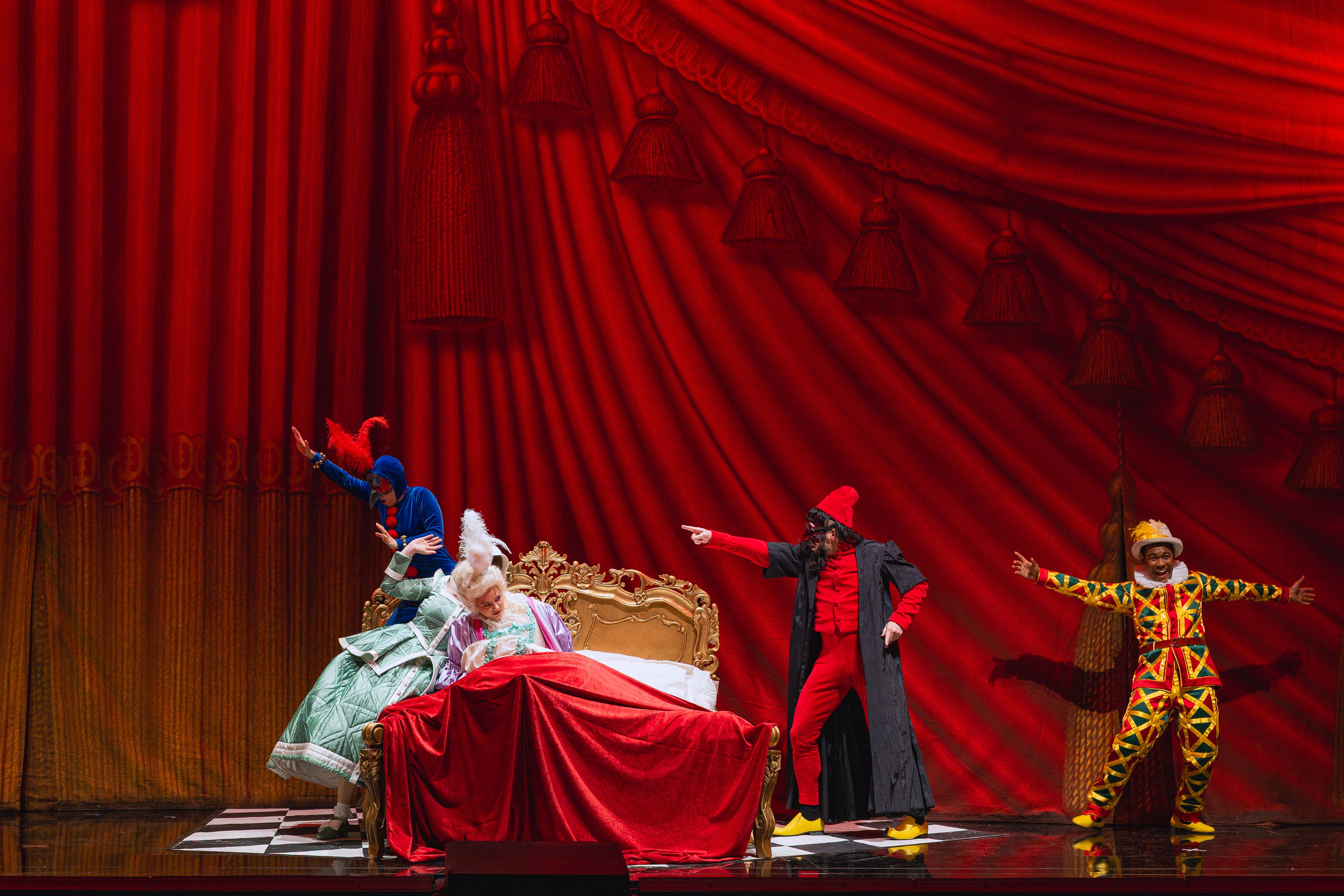 Das Szenefoto aus der Oper "Le nozze di Figaro zeigt vier Hauptdarsteller:innen in bunten Kostüme, die sich um ein barockes Bett mit roter Überdecke tummeln, vor tiefrotem Hintergrund, der wie ein Vorhang mit Falten und Quasten aussieht