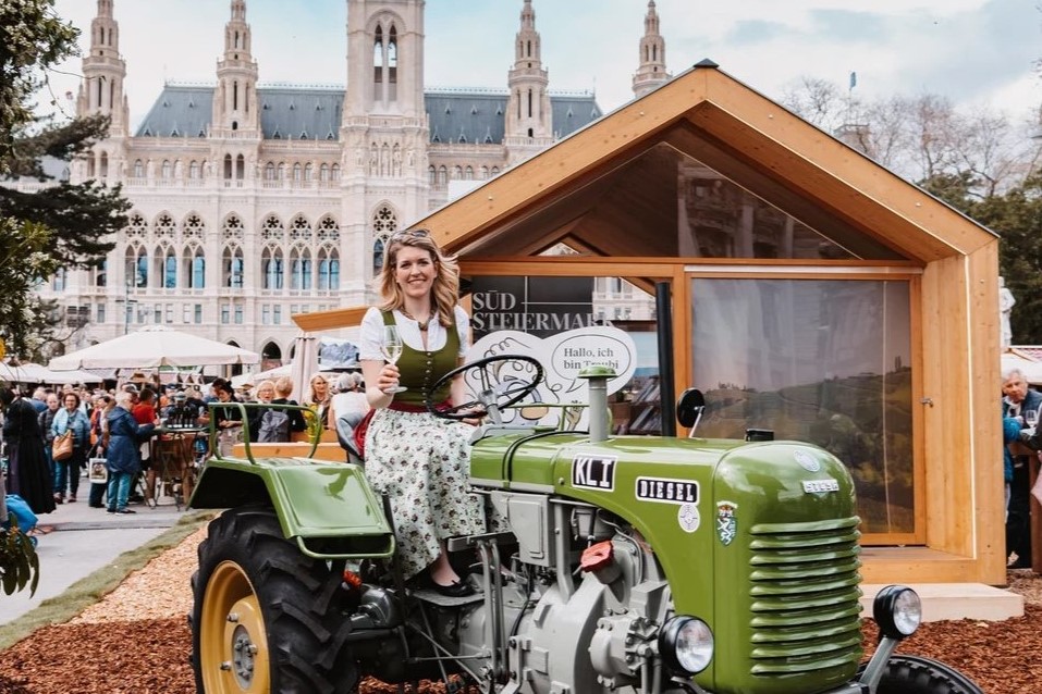Steiermark-Frühling vor dem Wiener Rathaus, im Vordergrund sitzt eine junge blonde Frau im Drindlkleid auf einem grünen Traktor