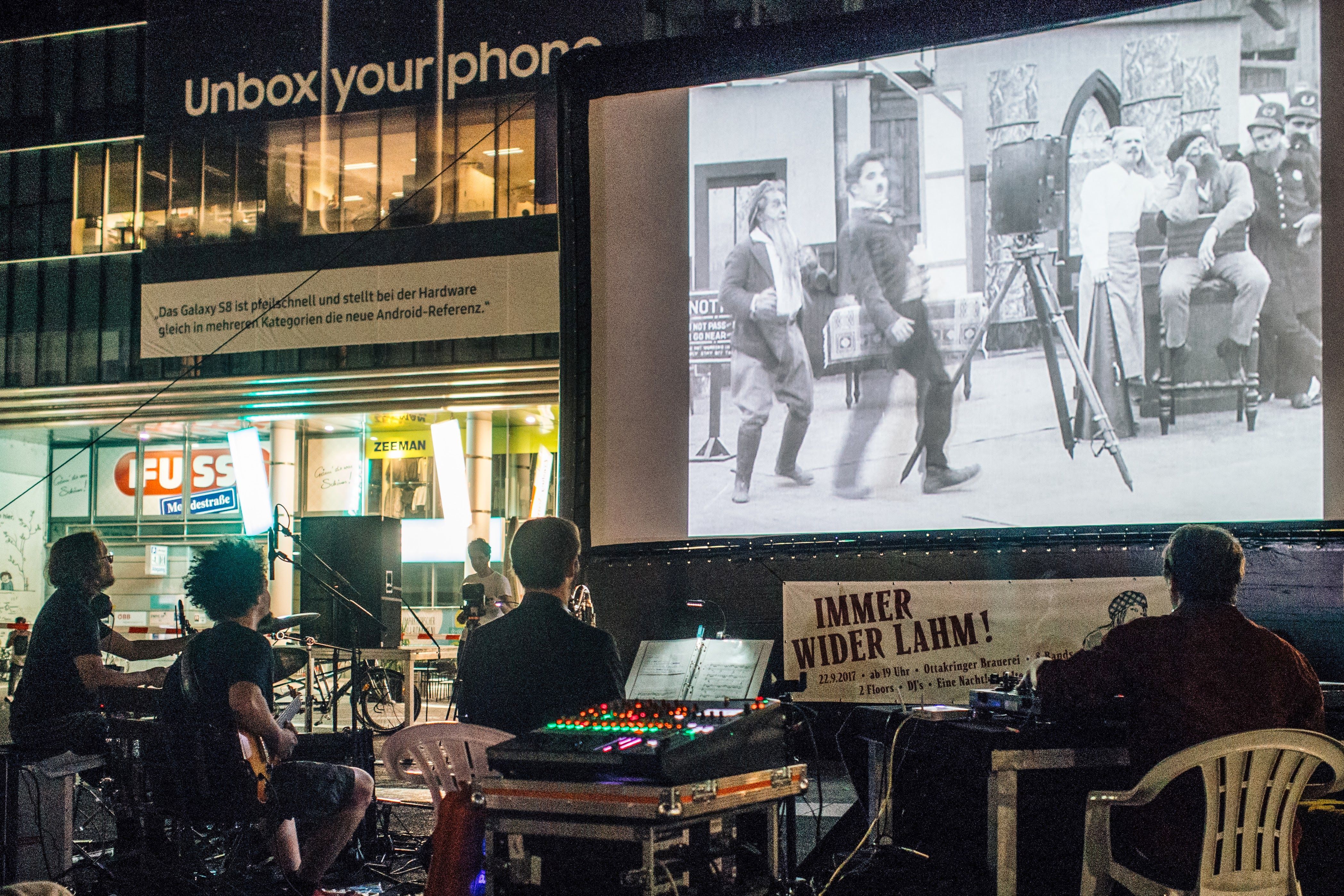 Photo showing screen with black and white film, in front of it musicians with instruments