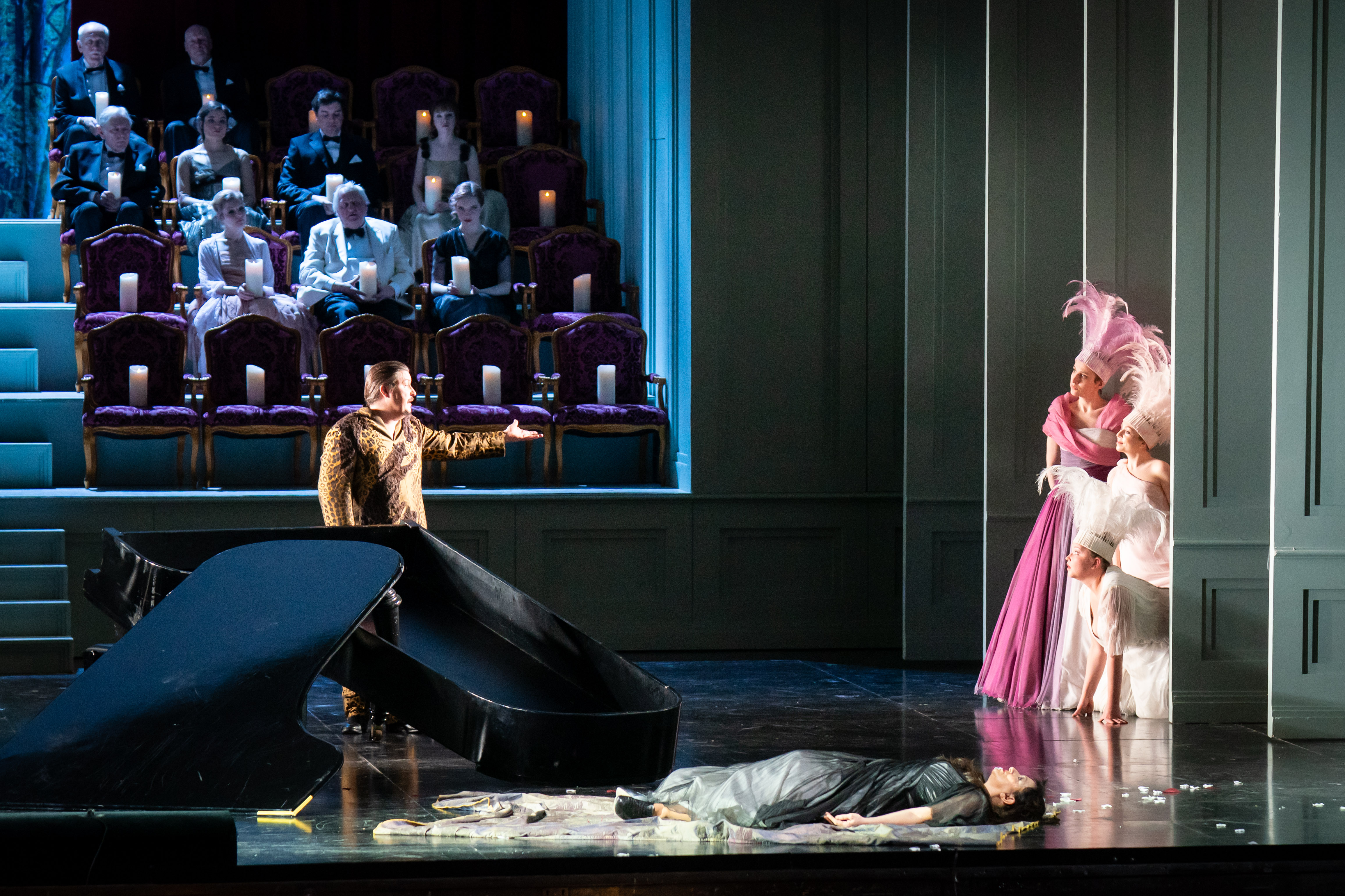 The scene photo from the opera “Ariadne auf Naxos” shows a woman in a gray costume lying on the floor in front of the shattered grand piano, behind the grand piano stands a man pointing to three women in pink to pale pink costumes with feather decorations