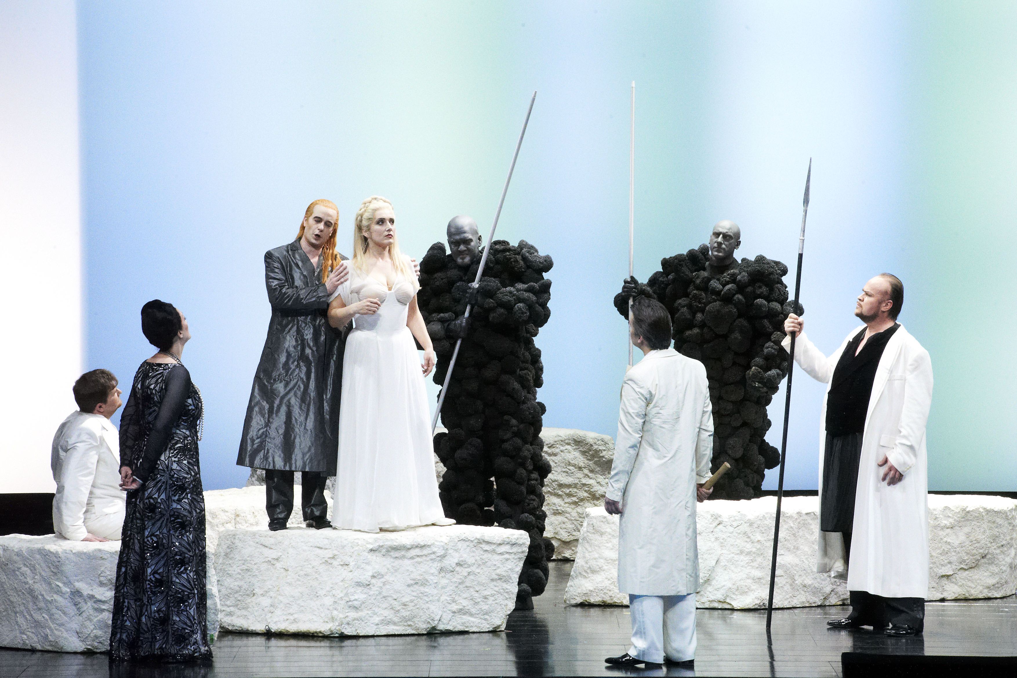 Das Szenefoto aus der Oper "Das Rheingold" aus dem Ring der Nibelungen zeigt die Sänger:innen in schwarzen und weißen Kostümen auf einer hellen Bühne. Im Zentrum eine blonde Frau in weißem Kleid, daneben ein Mann mit rotem langem Haar in schwarzem Mantel