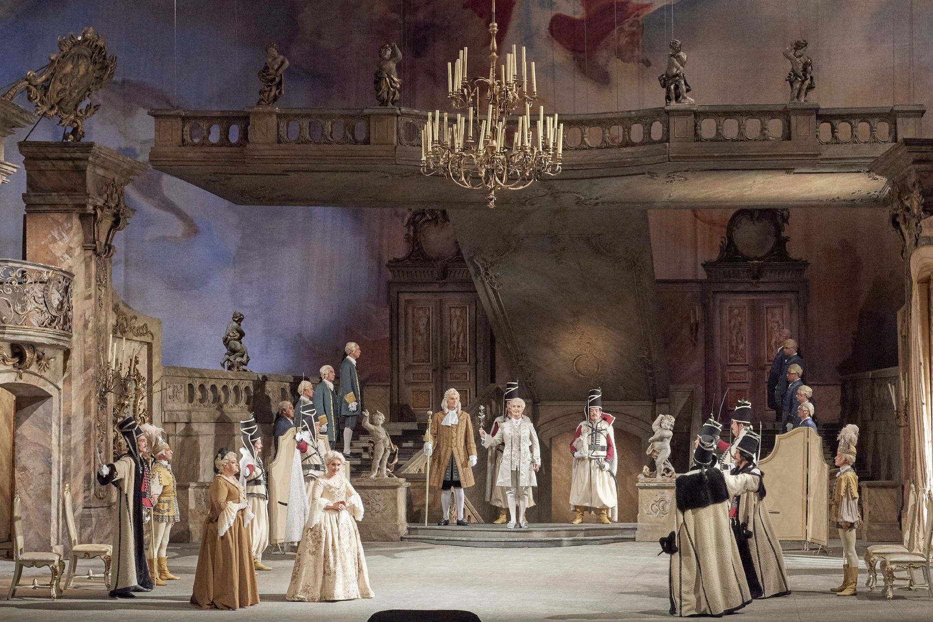 The scene photo from the opera “Der Rosenkavalier” of the Vienna State Opera shows a baroque hall in which numerous members of a court are located. The historical costumes and the hall are in shades of beige and brown.