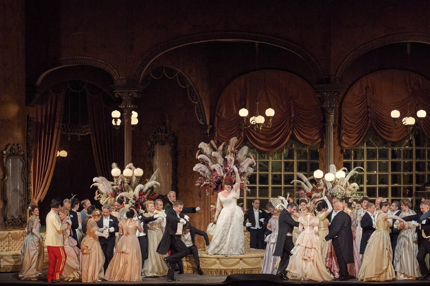 Scene photo from the operetta “Die Fledermaus”, showing an evening party in elegant dresses, tuxedos and uniforms in a festive setting, in the center the leading actress in a white evening dress standing on a pedestal
