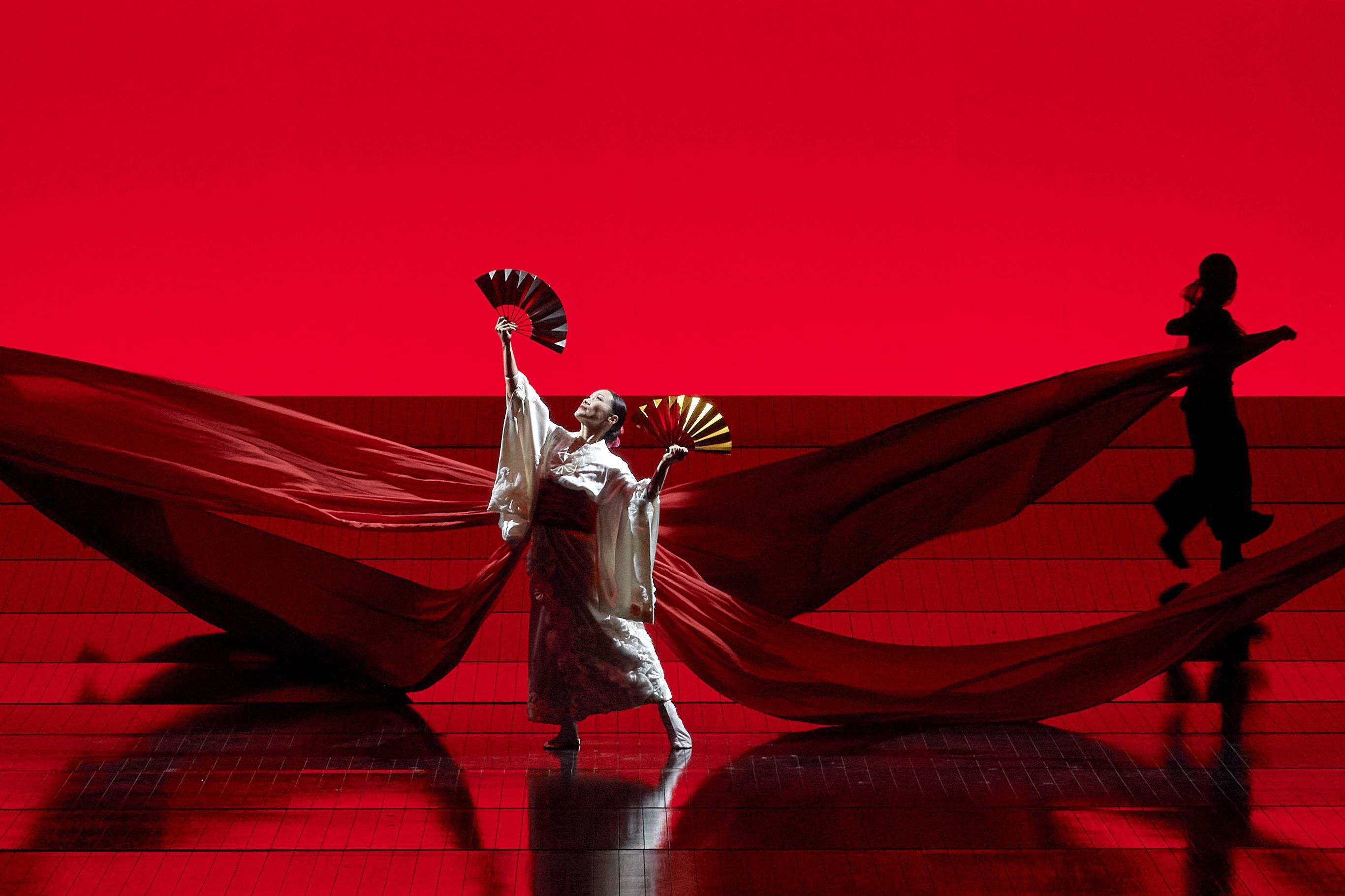 Scene photo of the opera Madama Butterfly, the leading actress in traditional Japanese dress with two fans in her hands in the spotlight against a deep red background