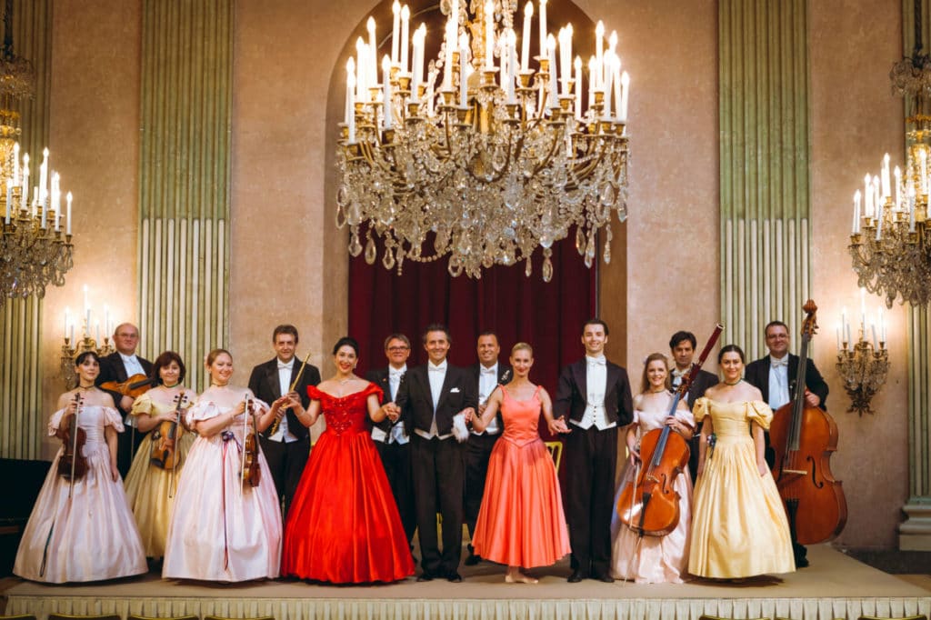  Photo of the Vienna Residence Orchestra, the members in festive evening dress and with instruments, in a historic hall with large chandeliers.