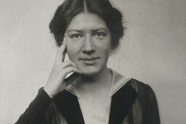 Zu sehen ist ein Porträtfoto in schwarzweiß von Käthe Leichter. Sie trägt ihr dunkeles Haar hochgesteckt, trägt ein elegantes Oberteil mit weißem Spitzenkragen 