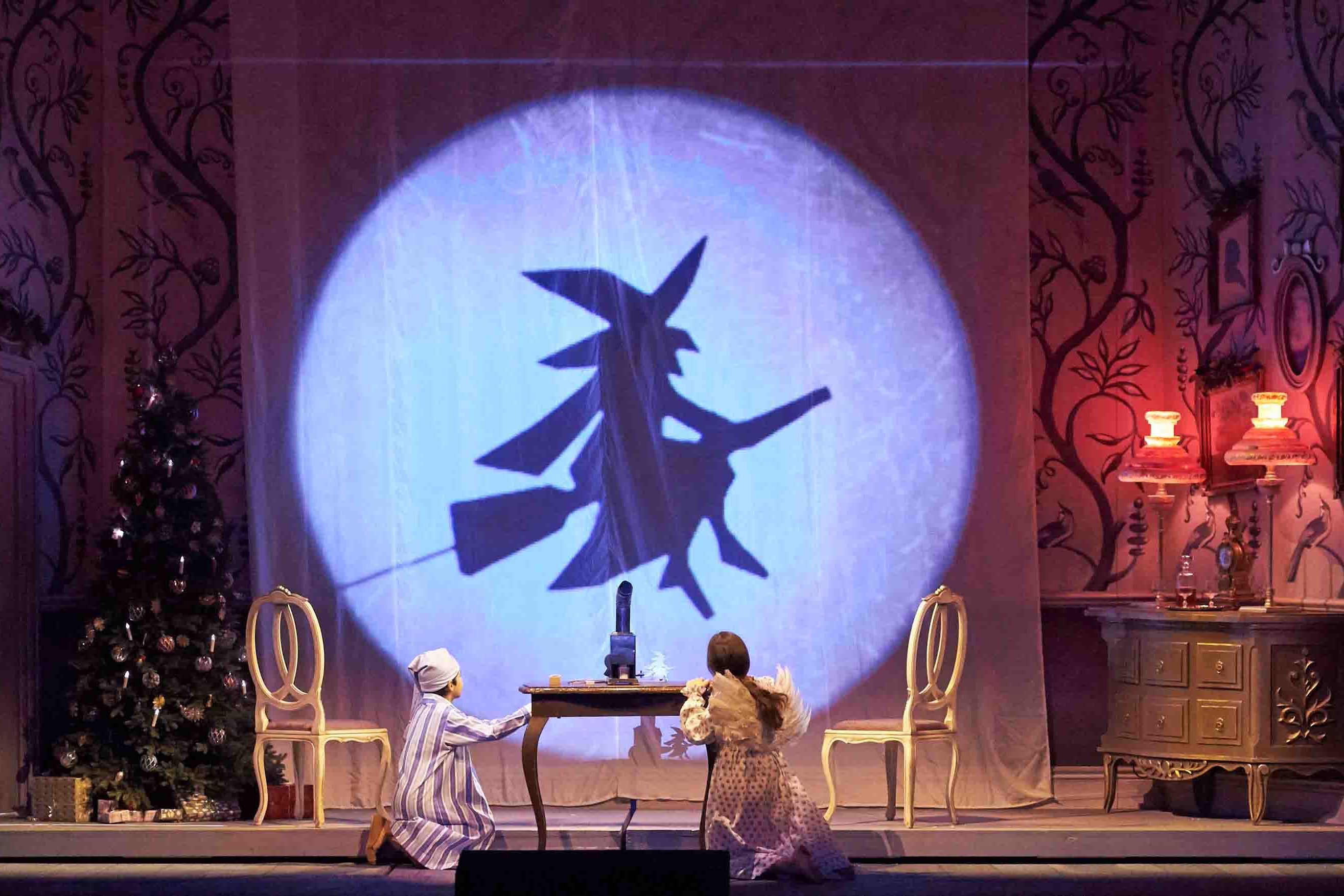 Eine Szene aus der Familienoper Hänsel und Gretel. Ein Bub und ein Mädchen in Nachtgewändern knien vor einem Tisch in einem historischen Kinderzimmer, rechts ein Weihnachtsbaum, im Hintergrund auf einer Leinwand die Silhouette einer Hexe auf einem Besen